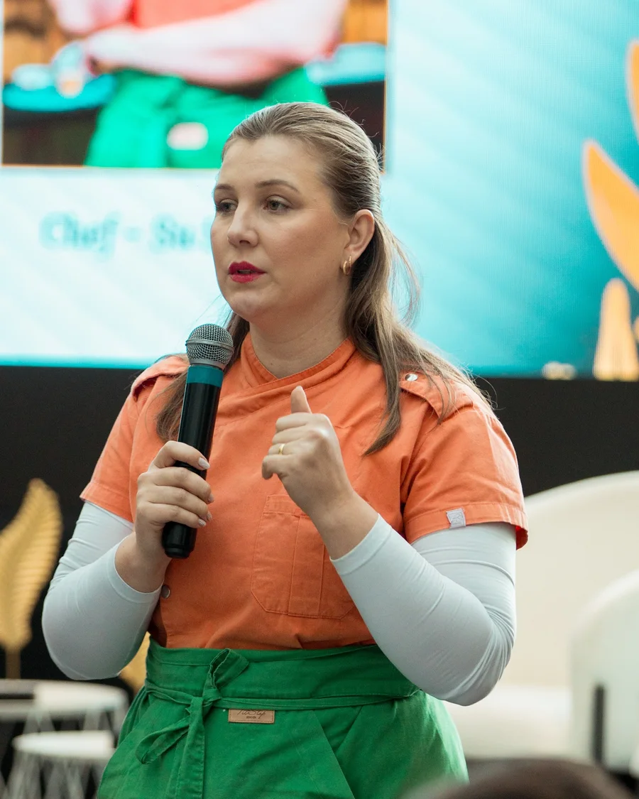 Su Selinger, chef da Sabores do Campo, falando ao microfone de dólmã laranja e avental verde